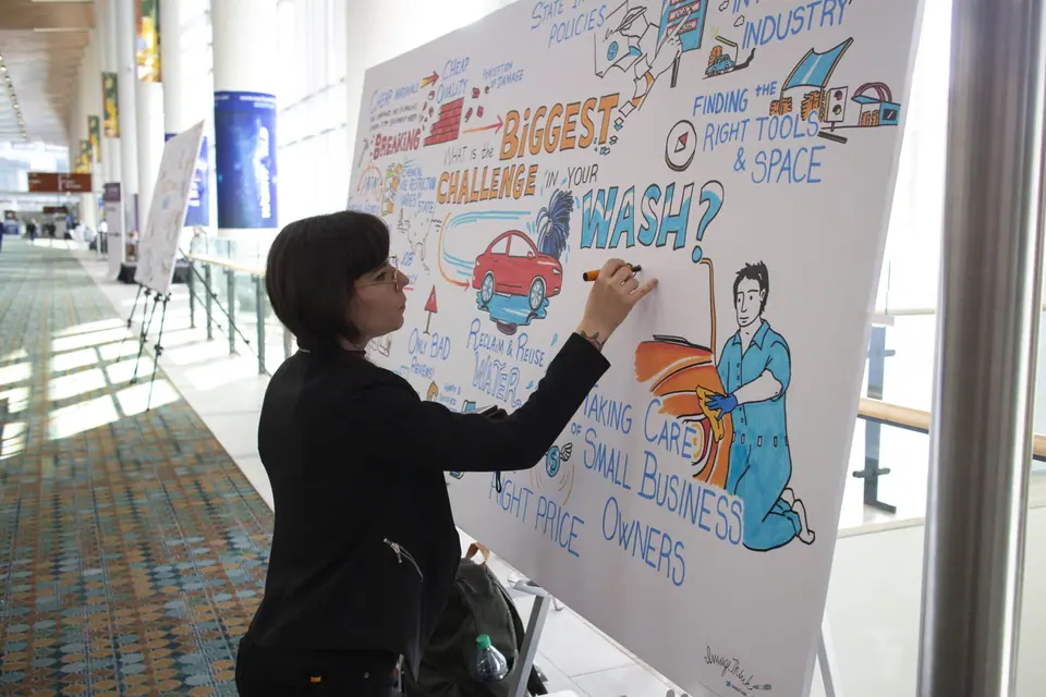 A graphic recorder from ImageThink draws live on a large foamcore board at a conference, capturing audience insights with colorful illustrations and text. The board features the central question “What is the biggest challenge in your wash?” surrounded by hand-drawn graphics of a red car, a man polishing a vehicle, and phrases about small business challenges like pricing, water reclamation, and industry tools. The artist, dressed in black, uses orange and blue markers while standing in a brightly lit convention hallway with patterned carpet and large windows. Other visual boards are visible in the background, showcasing a dynamic and creative conference environment.